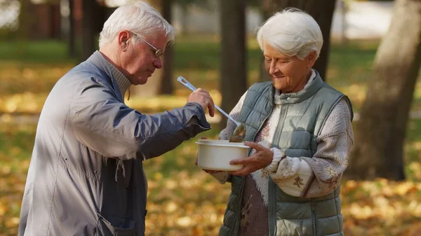 Genre caucasien retraité homme aux cheveux gris aide les personnes dans le besoin en partageant sa soupe maison avec une femme âgée sans abri. — Photo