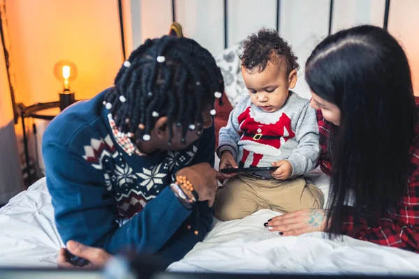 Gemengde race peuter krijgt een smartphone van zijn ouders terwijl ze op hun bed liggen. — Stockfoto