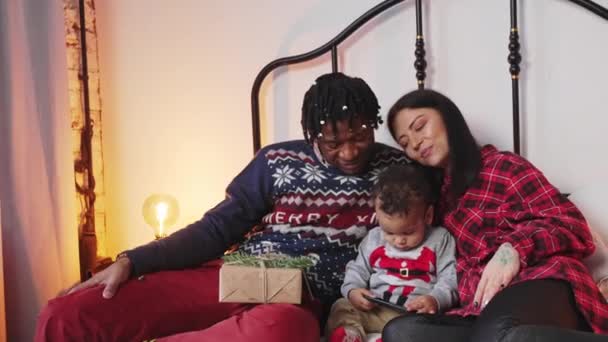 행복 한 혼합 인종 가족은 아기와 침대에 함께 앉아 크리스마스 시간을 보내고 있다. — 비디오