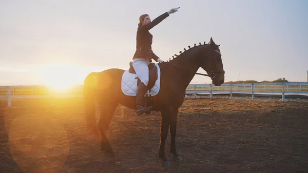 Uma jovem mulher bonita está sentada em um cavalo, e atrás dela está o pôr do sol e ela está apontando o dedo para o céu — Fotografia de Stock