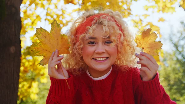 Glückliches junges Millenialmädchen in Rot mit blonden lockigen Haaren, das Herbstblätter hält - Porträt — Stockfoto