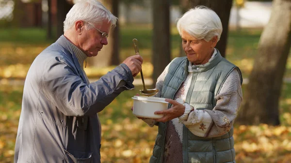 Generous elderly grey-haired caucasian man volunteers in charity organization and pours hot soup into the elderly homeless womans white bowl.