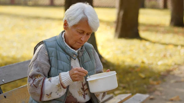Starsza, bezdomna, emerytowana, biała kobieta siedząca na ławce i jedząca ciepłą zupę, którą dała jej grupa ochotników.. — Zdjęcie stockowe