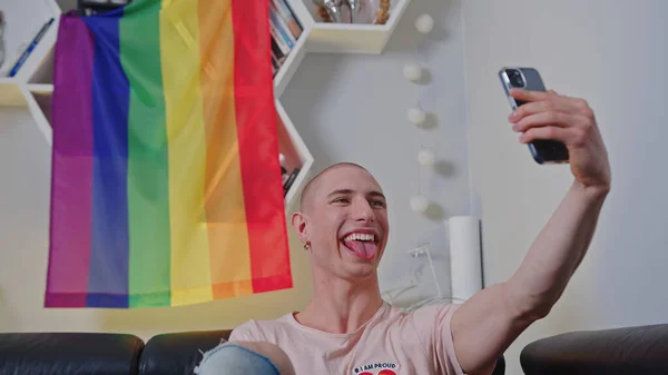 Gay Kaukasische kale gay man zitten in voor regenboog trots vlag en het nemen van selfie van zichzelf met zijn tong uit. — Stockfoto