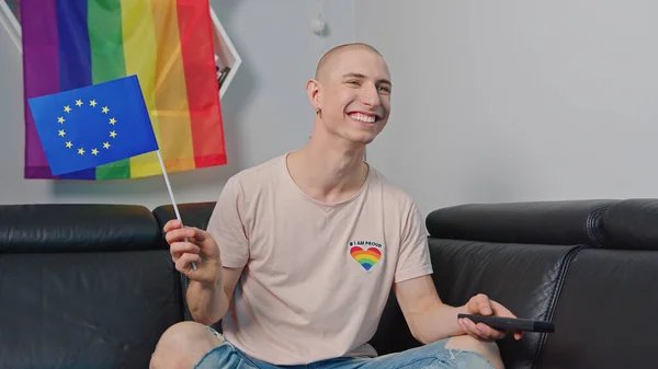 Caucásico europeo calvo gay hombre ondeando bandera europea, sentado en un sofá, sonriendo, y la celebración de control remoto. — Foto de Stock