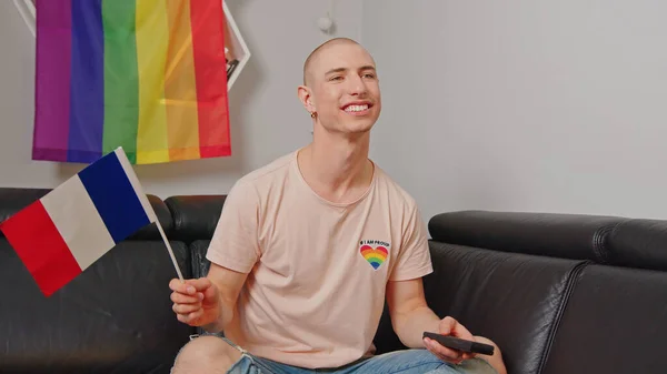 Francês gay homem que vive em Paris, acenando bandeira francesa, sentado em um sofá, e assistindo TV em seu apartamento. — Fotografia de Stock