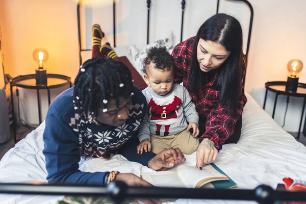 Karışık ırk evlilikleri yeni yürümeye başlayan çocuklarıyla yatağa uzanıp ona kitap okuyorlar.. — Stok fotoğraf