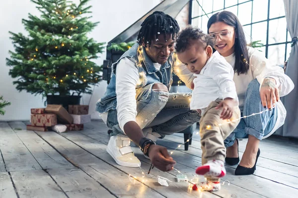 Χαρούμενη μικτή οικογένεια που παίζει με χριστουγεννιάτικα φώτα. Τα πρώτα Χριστούγεννα με ένα νήπιο. — Φωτογραφία Αρχείου