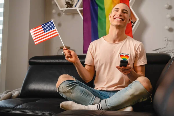 백인 미국인 여성 천 년 게이 남자 자신의 아파트에 검은 소파에 앉아 있는 동안 미국 국기를 흔들며. — 스톡 사진