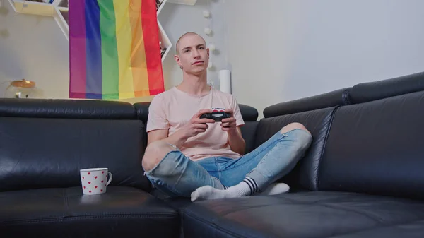 Ativista gay europeu sentado em um sofá e jogando videogames com seu gamepad. Arco-íris orgulho bandeira no fundo. — Fotografia de Stock