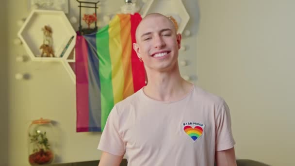 Persona queer no binaria señalando a su camiseta con corazón de arco iris. Tiro mediano interior con bandera de orgullo en el fondo. — Vídeos de Stock