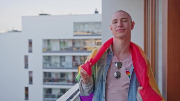 Dumny europejski millenijny gej stojący przed swoim mieszkaniem i przykrywający ramiona tęczową flagą dumy. — Wideo stockowe