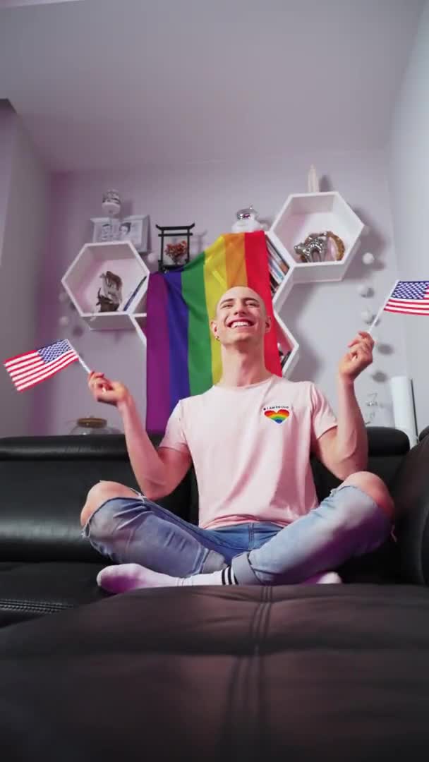 Amerykański aktywista gejowski walczący o prawa gejów, machający flagą USA siedząc na kanapie. Pionowe ujęcie wewnętrzne. — Wideo stockowe