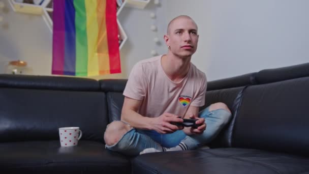 Caucasiano gay gamer homem jogar jogos com seu gamepad enquanto sentado no um preto sofá com arco-íris bandeira no o fundo. — Vídeo de Stock