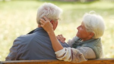 Kürsüde oturan yaşlı beyaz bir çift kocasının saçına dokunuyor. Arkasından çekilen gülümsüyor. 
