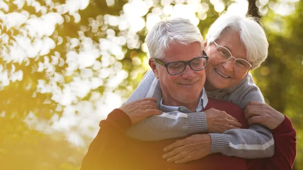 Retrato de feliz anciano caucásico pareja mujer abraza a su marido desde la espalda tanto sonriente hombre mirando hacia abajo, mujer mira hacia otro lado enfoque selectivo —  Fotos de Stock