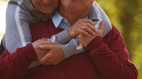Літня кавказька жінка обіймає свого чоловіка ззаду тримаючи один одного за руки портретний селективний фокус на руках без будь-яких облич. — стокове фото