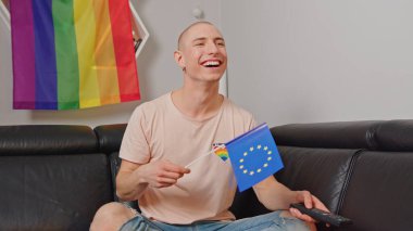 Kafkasyalı kel eşcinsel adam Avrupa bayrağı sallıyor, koltukta oturuyor, gülümsüyor ve uzaktan kumandayı elinde tutuyor..