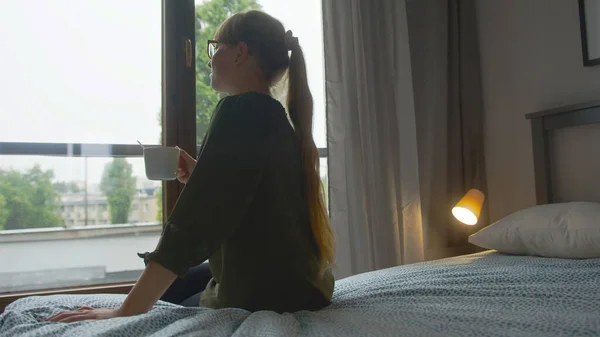 Foto nas costas de uma mulher caucasiana de cabelos compridos com 20 ou 30 anos sentada na cama em seu apartamento moderno e segurando uma caneca branca e olhando pela janela. — Fotografia de Stock