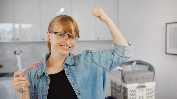 Mulher caucasiana independente em uma camisa jeans segurando uma ferramenta chave inglesa, olhando para a câmera com uma mão para cima, mostrando seus músculos. — Fotografia de Stock