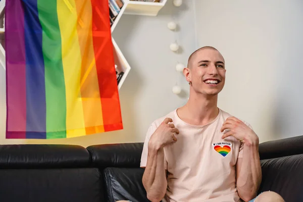 Bald queer caucasian 활동가는 소파에 앉아 TV 를 보고 손을 들고 미소짓고 있습니다. 뒤에 있는 무지개 깃발. — 스톡 사진