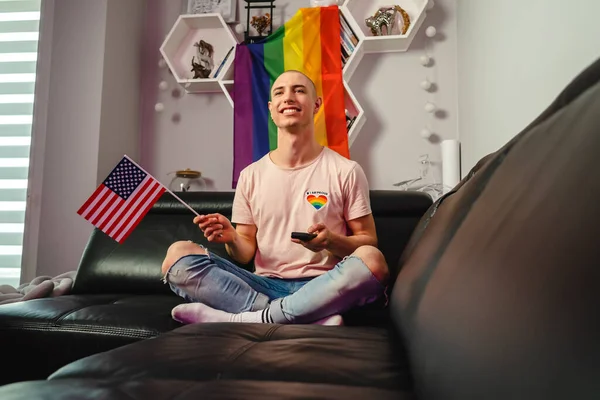 Caucásico americano queer millennial gay hombre ondeando USA bandera mientras sentado en un negro sofá y viendo tv en su apartamento. —  Fotos de Stock