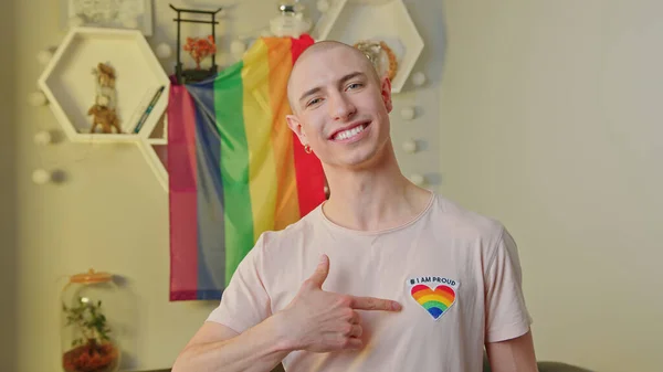 Heureux homme gay caucasien souriant à la caméra, pointant un cœur arc-en-ciel sur son t-shirt, posant dans son appartement, et debout devant le drapeau de fierté arc-en-ciel. — Photo