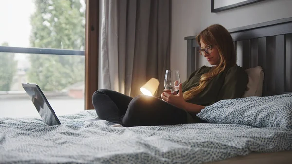 Concepto de tiempo libre solo. Mujer caucásica de pelo largo de mediana edad relajándose en su cama, sosteniendo una copa de vino y viendo una película en su computadora portátil. — Foto de Stock