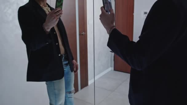 Bare borst vet Kaukasische queer persoon in een zwarte blazer en hoge hak schoenen kijken naar zichzelf in de spiegel en het nemen van een selfie., — Stockvideo