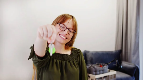 Begeisterte europäische Ingwerhaarige lächelt und zeigt Schlüssel zur Kamera, während sie in ihrem Wohnzimmer steht. — Stockfoto