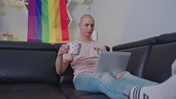 Europeiska queer person i 20-årsåldern sitter på en svart soffa, med bärbar dator, och dricka te eller kaffe från en stor mugg. — Stockvideo