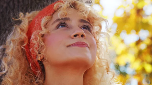 Positieve energie onder jonge volwassenen. Mooi blond meisje met krullend haar en rode hoofdband naar boven kijkend terwijl ze onder een boom zit. — Stockfoto