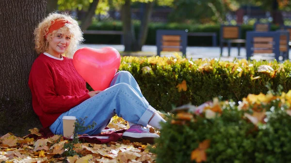 Junge blonde Europäerin sitzt unter dem Baum im Park und hält einen Luftballon in Herzform im Hintergrund — Stockfoto