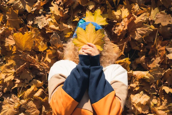 Herbstliches Glück. Junges kaukasisches Teenager-Mädchen legt sich auf den Rücken auf trockene, gelbe Blätter und bedeckt ihr Gesicht mit einem Blatt. — Stockfoto