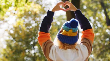 Sarışın, kıvırcık saçlı, elleri havada, kalp işareti gösteren birinin sırtından çekilmiş bir fotoğraf. Güzel güneşli hava, sonbahar titreşimleri.