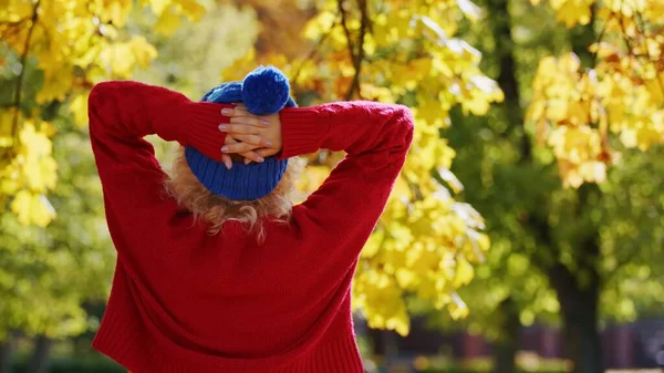 Laid-back caucasian young person standing backwards to the camera in red sweater and blue winter hat. Autumn colors trees in the background. — Stockfoto