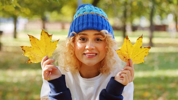 Cute European blonde curly-haired girl in a warm blue hat looking at camera and smiling while holding yellow leaves between her fingers. Autumn vibes. Outdoor shot. — Stockfoto