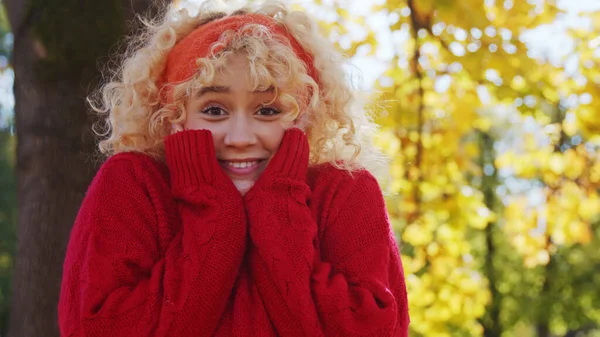 Aufgeregtes junges kaukasisches Mädchen in rotem Strickpullover, das in die Kamera blickt und ihr Smiley-Gesicht berührt. Schuss ins Freie. Herbstblätter im Hintergrund. — Stockfoto