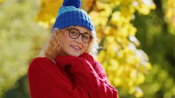 Outdoor autumn portrait of a cosily dressed caucasian blond girl looking at camera and smiling. Warm sweater and knitted blue hat. — Stockfoto