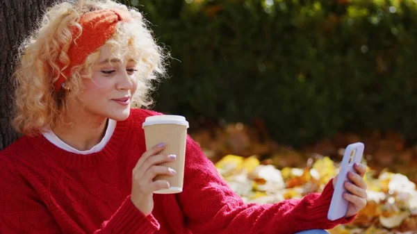 Cute caucasian girl with beautiful short curly blonde hair talking a selfie in a park while holding a cup of takeaway coffee or tea. Autumn scenery. — стоковое фото