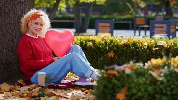 Beautiful curly-haired blonde caucasian girl holding heart-shaped red balloon and looking to the camera while sitting on autumn leaves under a tree. — Stockfoto