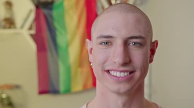 Kameraya bakıp göz kırpan, güler yüzlü, beyaz, kel, eşcinsel bir adamın yakın plan çekimi. Arka planda gökkuşağı bayrağı. Yüksek kalite 4k görüntü