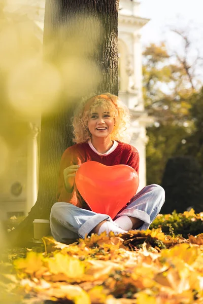 Attractive blond curly haired girl holds red balloon in the form of heart looking away and laughs full shot copy space autumn park background — Stockfoto