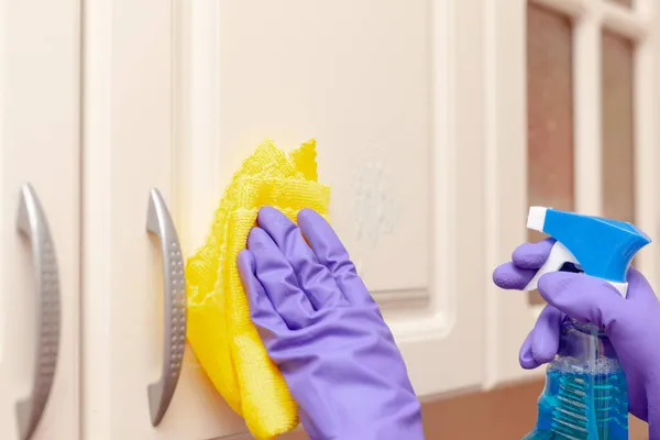 Mano femenina en un guante de goma púrpura limpia la puerta del gabinete de cocina beige. Trapo amarillo en la mano — Foto de Stock