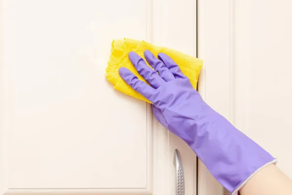 Mano femenina en un guante de goma púrpura limpia la puerta del gabinete de cocina beige. Trapo amarillo en la mano — Foto de Stock