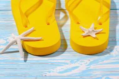 Sarı kauçuk plaj terlikleri ve mavi ahşap arka planda deniz yıldızı, metnin altına.