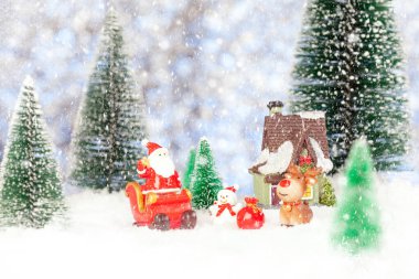 Yeni yıl tebrik kartı. Oyuncak bir Noel Baba Noel kızağının üstünde duruyor. Yakınlarda bir oyuncak ev, bir kardan adam, bir geyik var. Kar yağıyor. Oyuncak köknar ağaçları