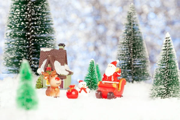 New year greeting postcard. A toy santa claus stands on a Christmas sleigh. Nearby there is a toy house, a snowman, a deer. Snow and toy firs all around — Stock Photo, Image