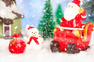 Yeni yıl tebrik kartı. Oyuncak bir Noel Baba Noel kızağının üstünde duruyor. Yakınlarda bir oyuncak ev, bir kardan adam, bir geyik var. Kar yağıyor. Oyuncak köknar ağaçları