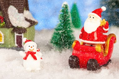 Yeni yıl tebrik kartı. Oyuncak bir Noel Baba Noel kızağının üstünde duruyor. Yakınlarda bir oyuncak ev var, bir kardan adam. Kar yağıyor. Oyuncak köknar ağaçları